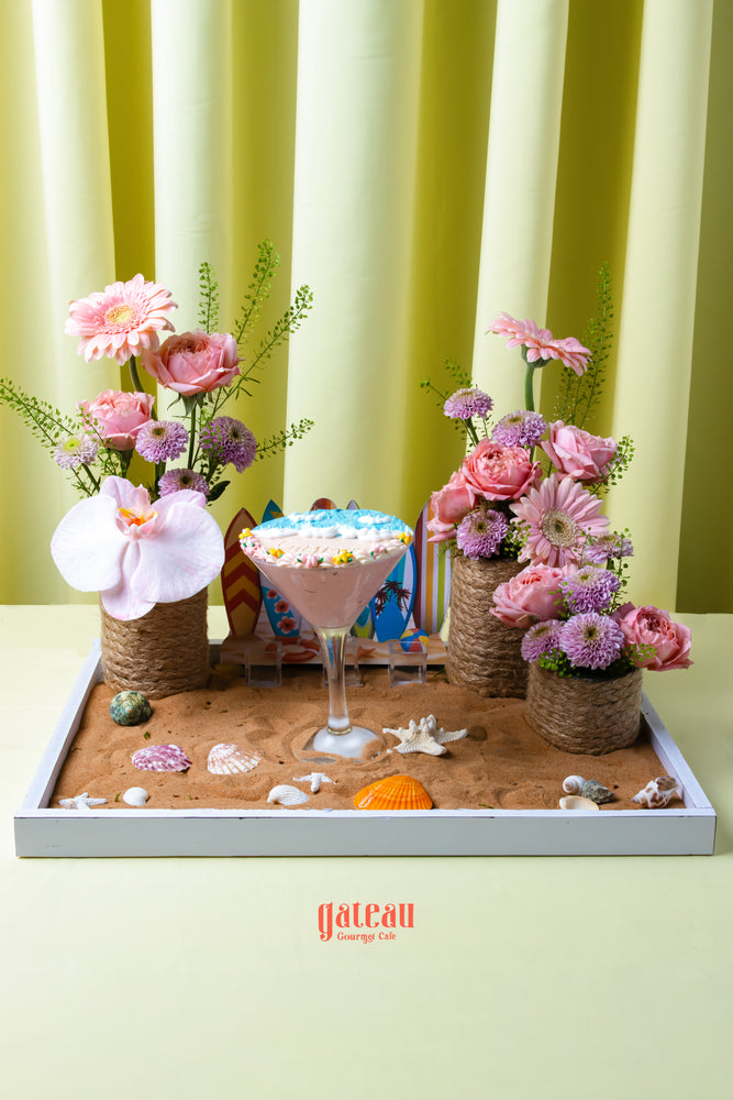 Floral beach cake dessert by Gateau Gourmet Cafe featuring pink flowers and seashell decorations on sand