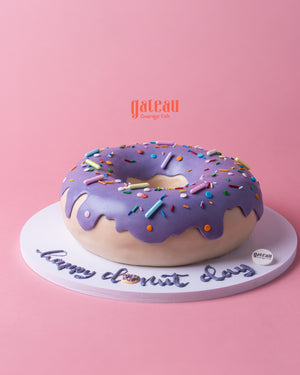 Gateau Gourmet Cafe custom donut-shaped cake with purple frosting and colorful sprinkles on a white base