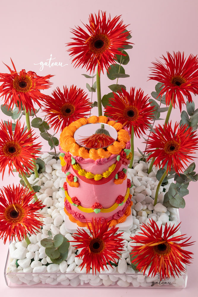 Custom floral cake design by Gateau Gourmet Cafe featuring vibrant pink and orange icing with surrounding red flowers