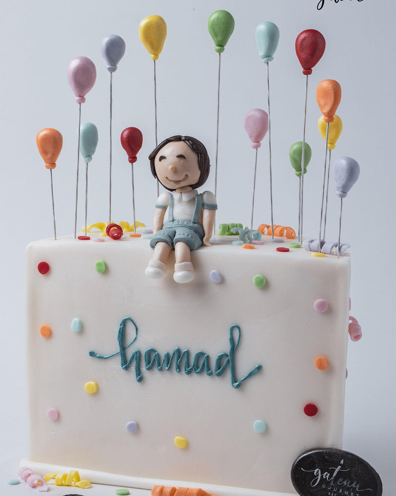 Custom pastel theme cake with a fondant boy figurine and colorful balloon decorations by Gateau Gourmet Cafe