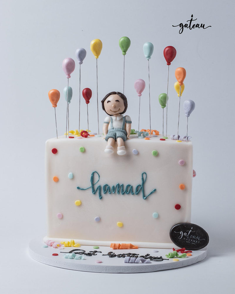 Custom pastel theme cake by Gateau Gourmet Cafe featuring a fondant child figure and colorful balloon decorations