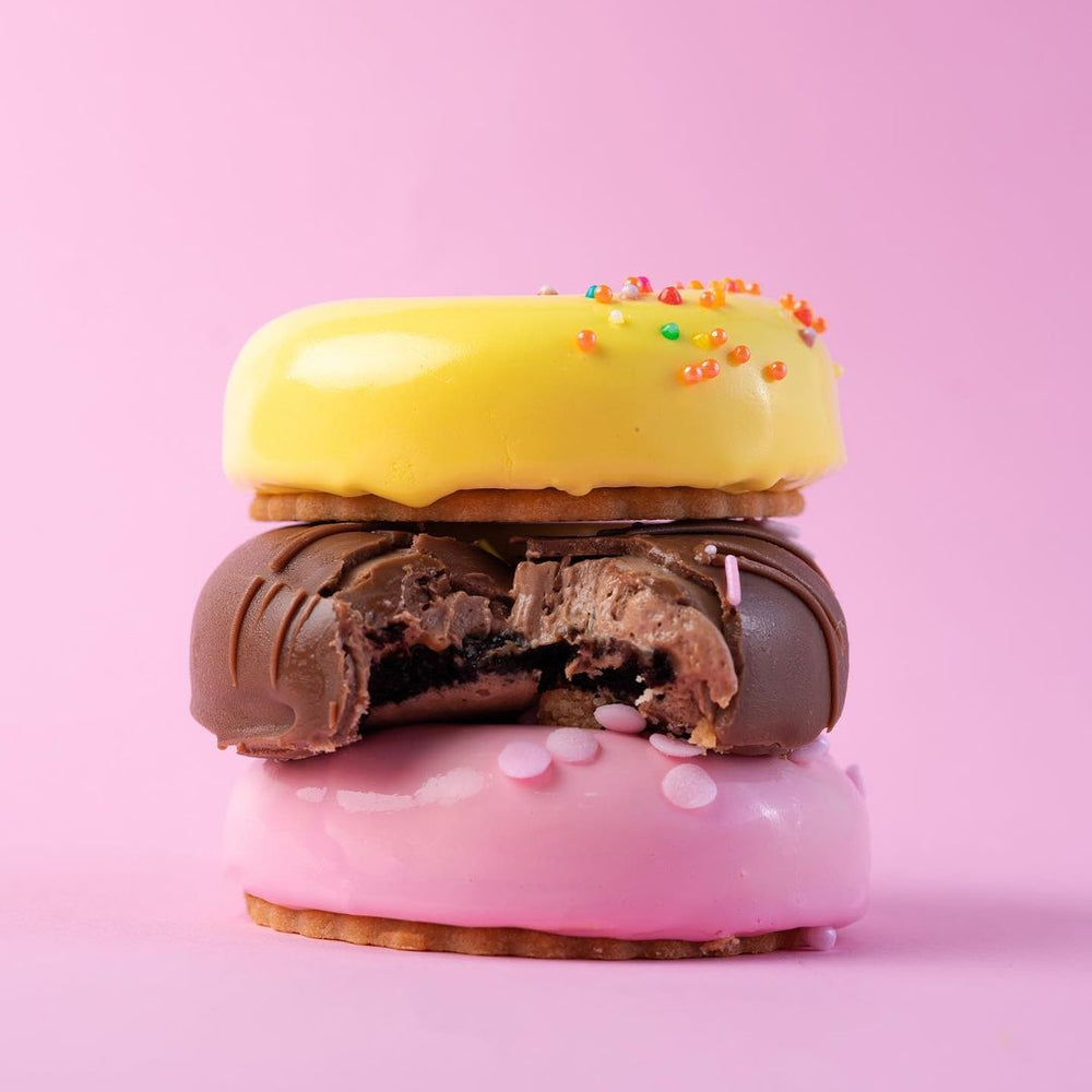 Gateau Gourmet Cafe colorful donut mousse sticks stacked with yellow, chocolate, and pink glaze on a pink background