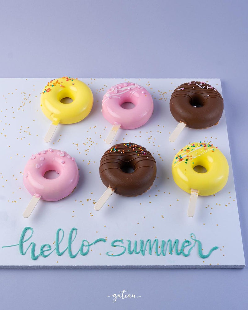 Colorful donut mousse sticks with yellow, pink, and chocolate coatings on sticks arranged on a white board