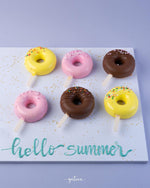 Colorful donut mousse sticks with yellow, pink, and chocolate coatings on sticks arranged on a white board