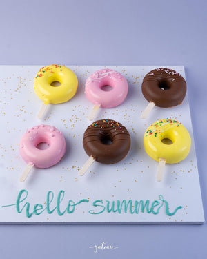 Colorful donut mousse sticks with yellow, pink, and chocolate coatings on sticks arranged on a white board