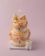 Fionka celebration cake by Gateau Gourmet Cafe with two tiers decorated in peach and white frosting with a bow and teddy bear