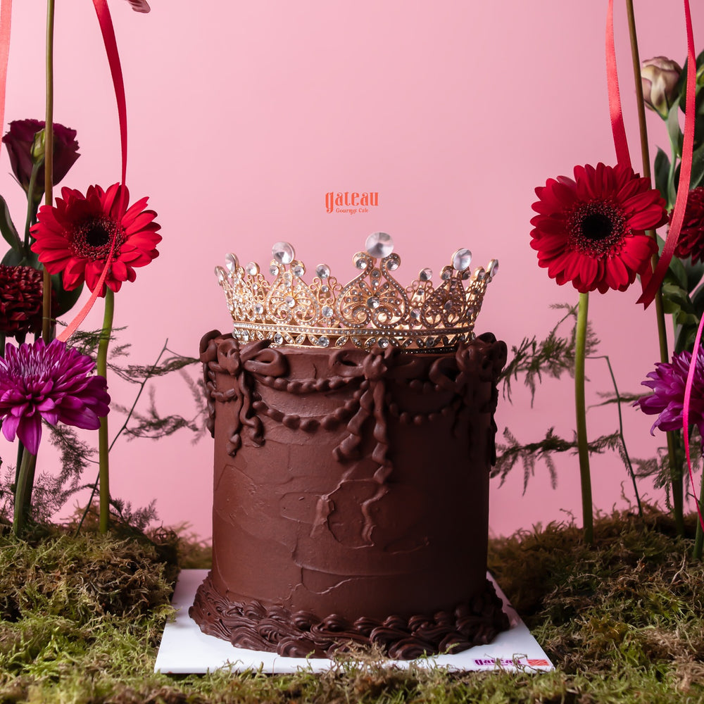 Gateau Gourmet Cafe chocolate cake with floral cake decor and a jeweled crown topper on pink background