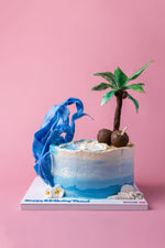 Beach theme cake by Gateau Gourmet Cafe with palm tree, coconuts, and blue wave decoration on white base