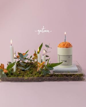 Birthday cake with flower arrangement featuring orange frosting and green foliage on a moss tray