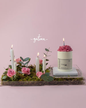 Birthday cake with flower arrangement featuring pink roses, candles, and greenery on a clear tray