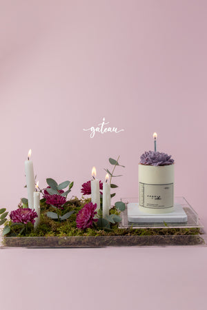 Birthday cake with flower arrangement featuring purple flowers and candles on a decorative tray by Gateau Gourmet Cafe