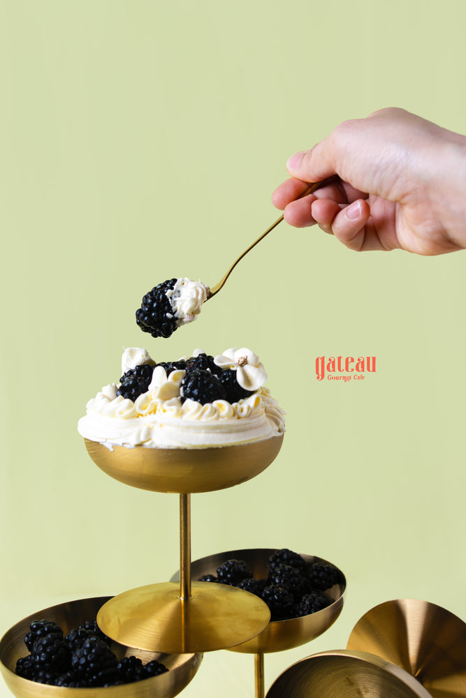 Hand holding a spoon above a gold dessert cup filled with blackberry dessert cup topped with cream and fresh blackberries