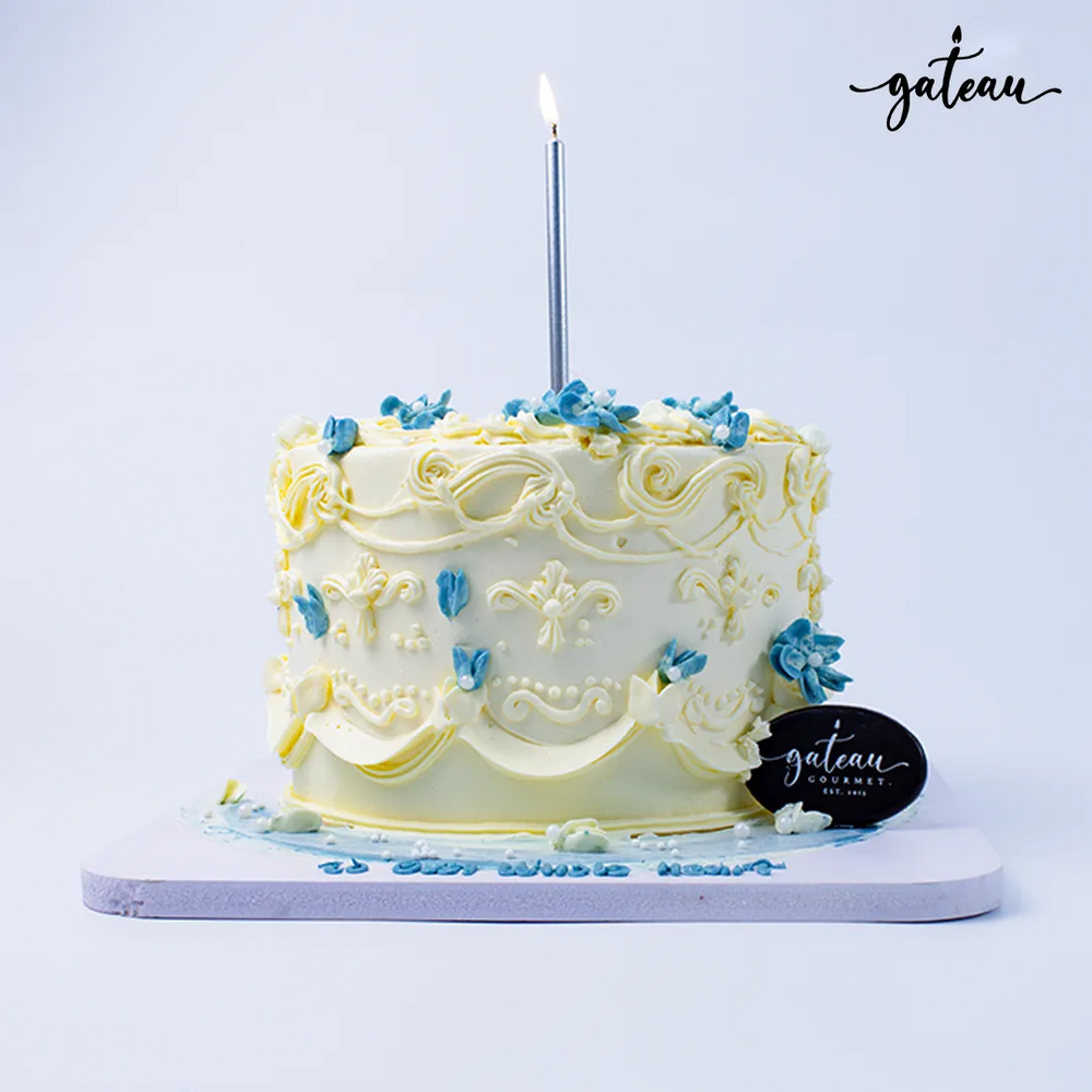 Gateau Gourmet Cafe buttercream celebration cake with yellow and blue floral decorations and a single silver candle