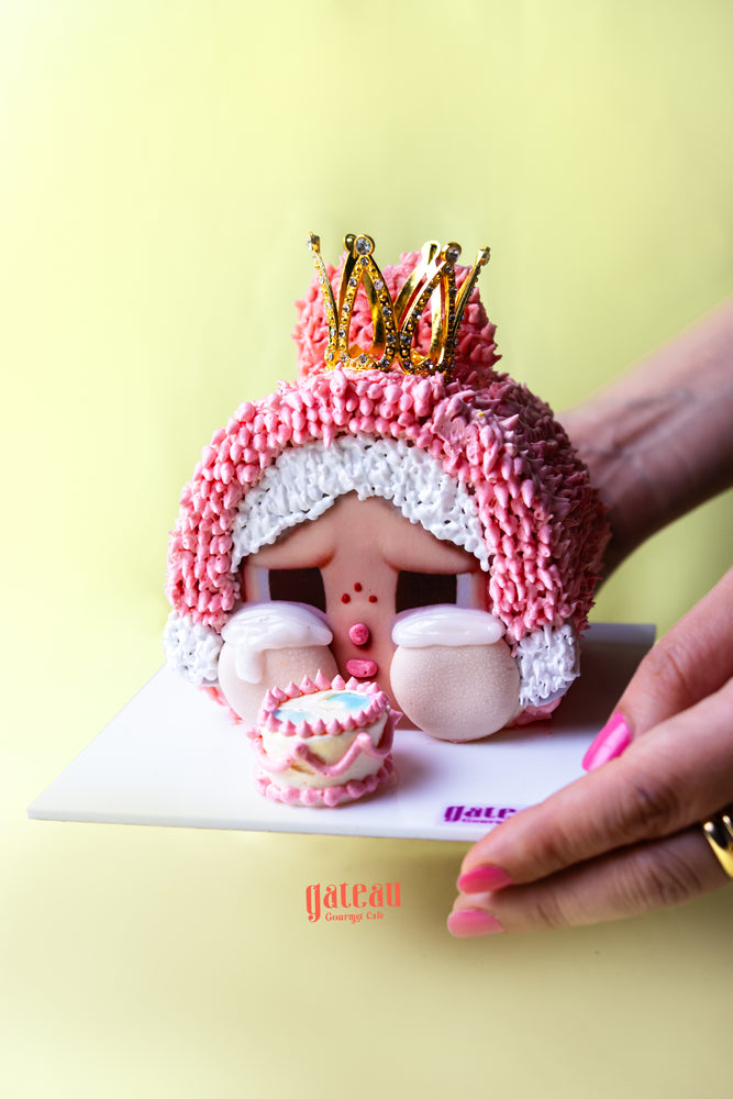Gateau Gourmet Cafe CRYBABY character cake with pink frosting and gold crown on white serving board