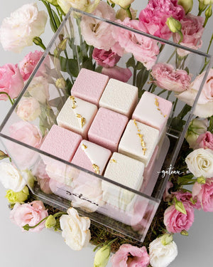 Cube cake gift box with pink and white decorated cakes inside a clear box surrounded by flowers