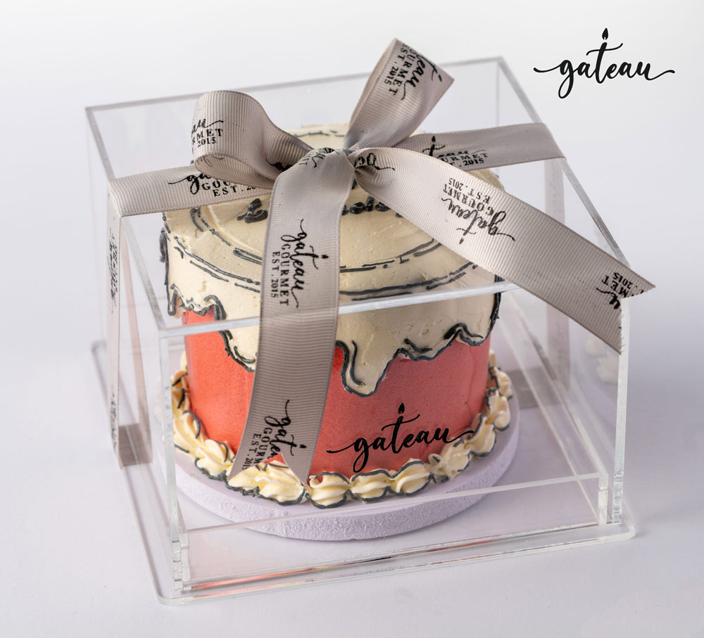 Custom colored cakes displayed in a clear box with elegant ribbon from Gateau Gourmet Cafe