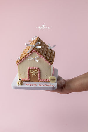 Gateau Gourmet Cafe custom house-shaped cake with pink and beige frosting held on hand against pink background