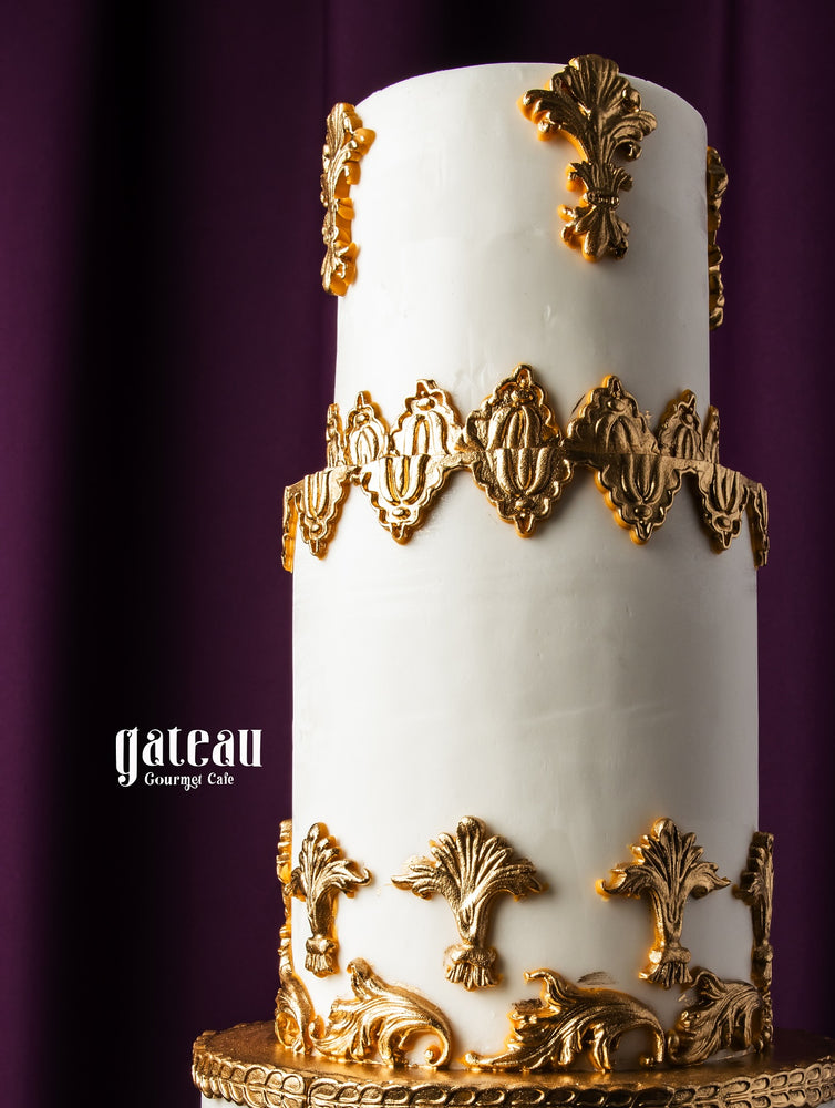 Gateau Gourmet Cafe custom royal cake with white icing and intricate gold decorative accents on a three-tier design