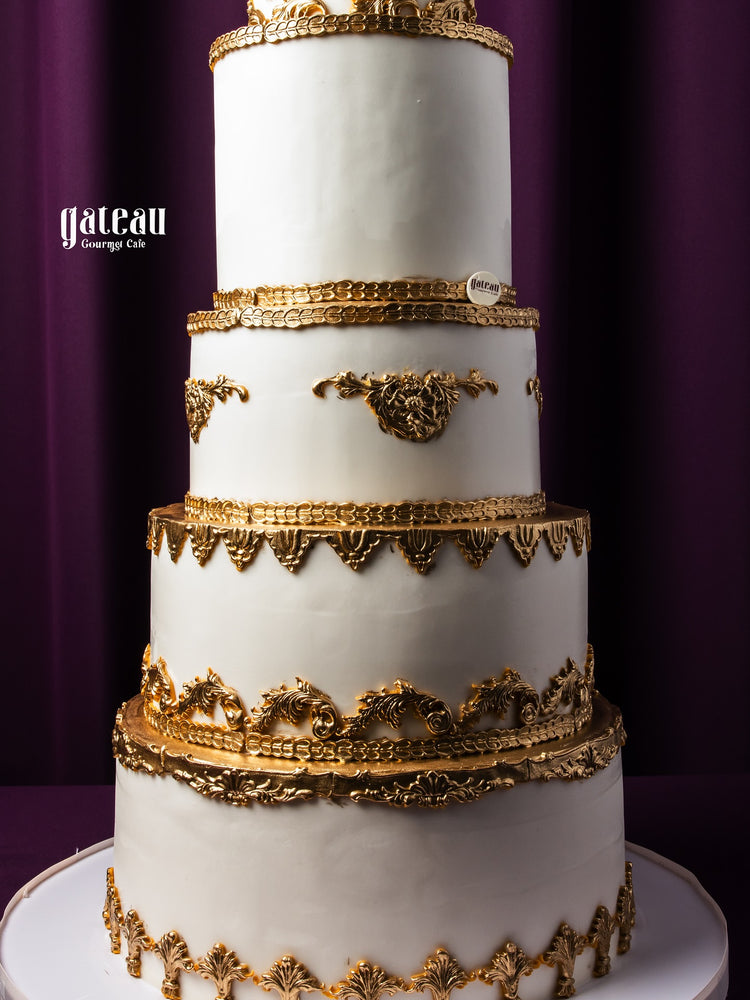 Elegant four-tier custom royal cake with white fondant and intricate gold decorations by Gateau Gourmet Cafe