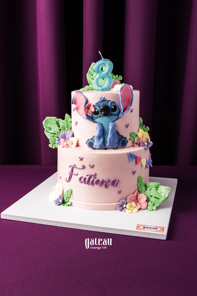 Two-tier custom Stitch themed cake with pink icing and colorful floral decorations by Gateau Gourmet Cafe