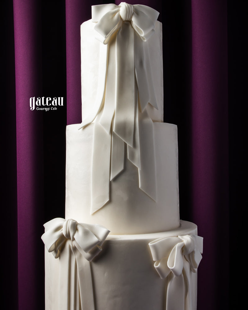 Elegant white three-tier custom wedding cakes with large decorative bows by Gateau Gourmet Cafe