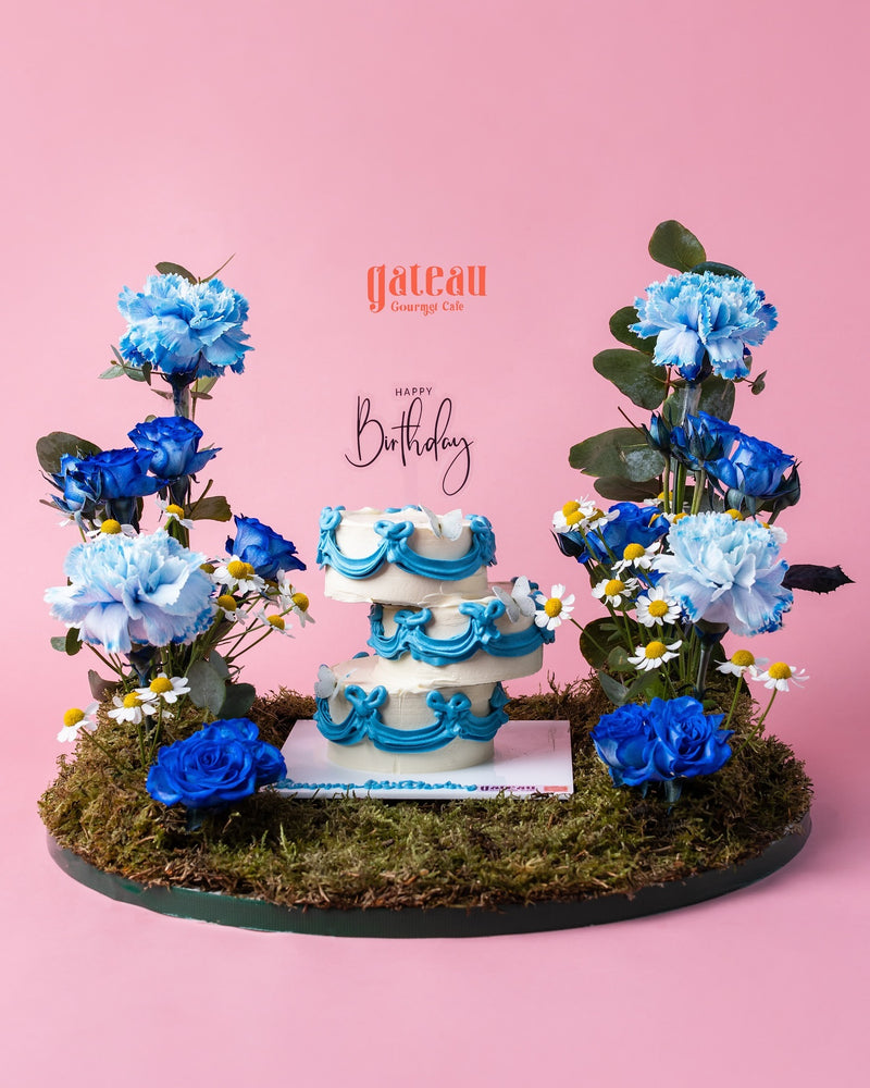 Decorative cake arrangement with blue and white floral accents on moss base from Gateau Gourmet Cafe