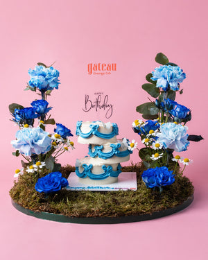 Decorative cake arrangement with blue and white floral accents on moss base from Gateau Gourmet Cafe