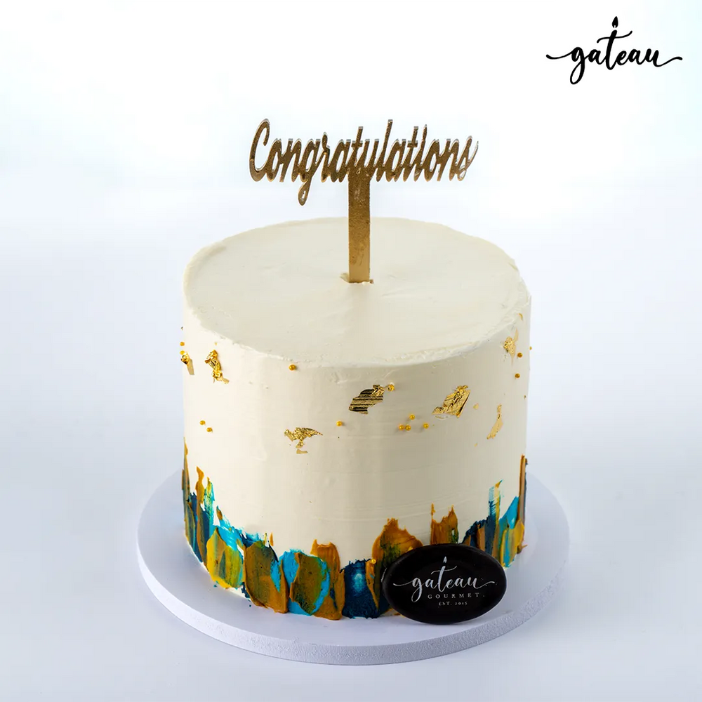 Decorative celebration cake with white frosting, gold accents, and congratulations topper by Gateau Gourmet Cafe