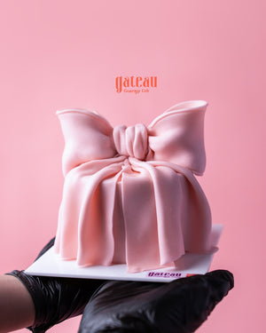 Pink Fionka Cake with elegant bow design held by hands wearing black gloves from Gateau Gourmet Cafe