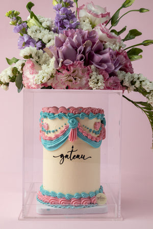 Floral cake arrangement featuring a tall pastel pink and blue decorated cake with fresh purple and white flowers on top