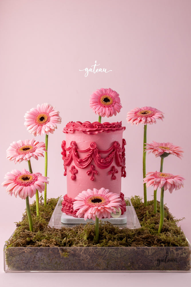 Pink cake with intricate pink floral cake decorations surrounded by fresh pink flowers on moss base