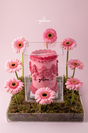 Pink cake with intricate floral cake decorations surrounded by pink gerbera daisies on moss base