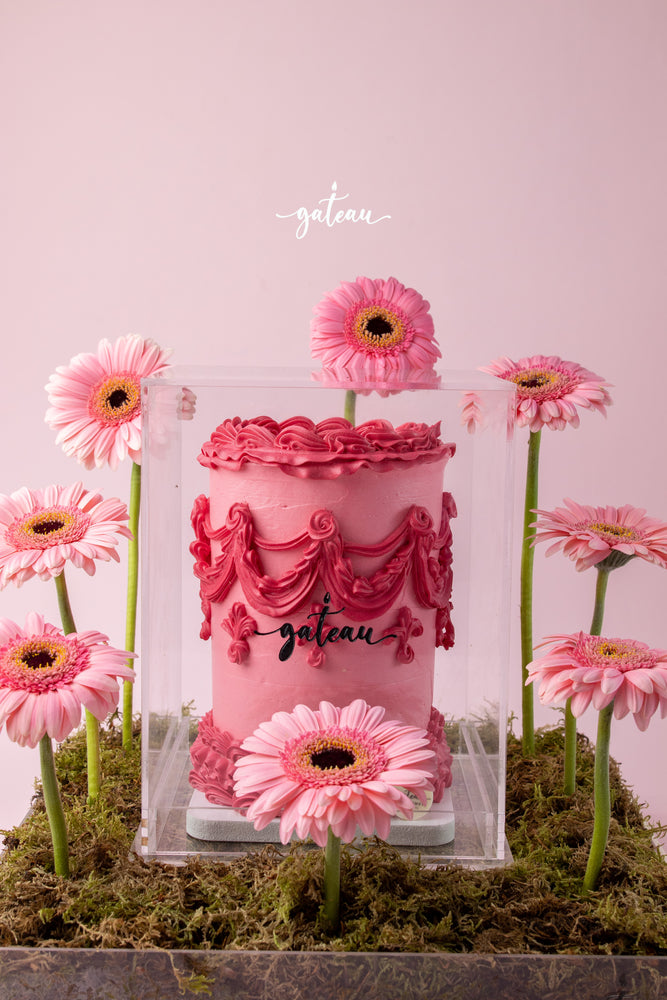 Gateau Gourmet Cafe pink cake with intricate floral cake decorations surrounded by pink flowers in clear display case
