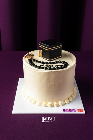 Hajj celebration cake by Gateau Gourmet Cafe featuring a white frosted cake topped with a black Kaaba replica and prayer beads