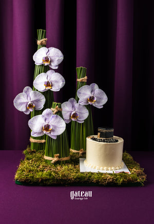 Gateau Gourmet Cafe Hajj celebration cake with white frosting and black decoration beside orchid floral arrangement on moss base