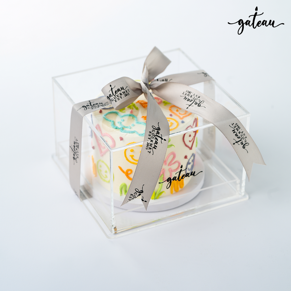 Hippie sticker cake displayed in a clear box with a ribbon from Gateau Gourmet Cafe
