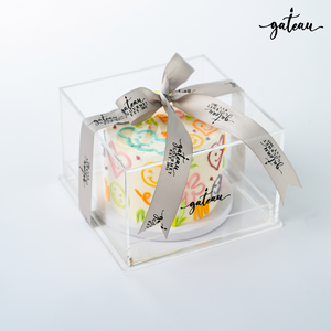 Hippie sticker cake displayed in a clear box with a ribbon from Gateau Gourmet Cafe