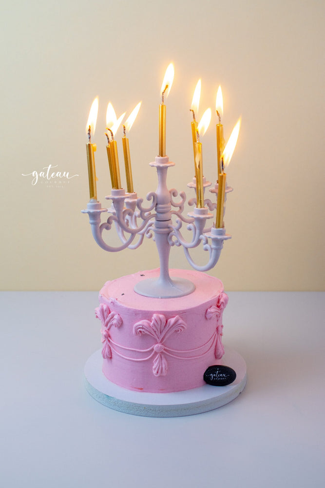 Pink cake from Gateau Gourmet Cafe decorated with a white luxury candelabra cake topper with lit golden candles
