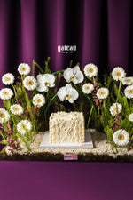 Luxury floral cake display by Gateau Gourmet Cafe featuring a decorated square cake surrounded by white flowers