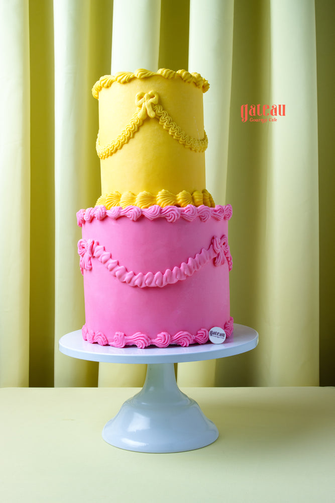Two-tier yellow and pink cake with decorative icing on white stand by Gateau Gourmet Cafe order gourmet cakes online