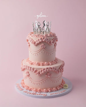 Two-tiered pink royal cake design with intricate icing decorations and silver crown topper from Gateau Gourmet Cafe