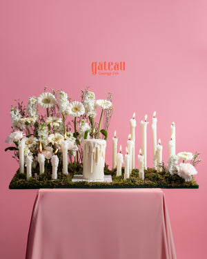 Romantic white cake and candle surrounded by white flowers on a moss base with a pink backdrop