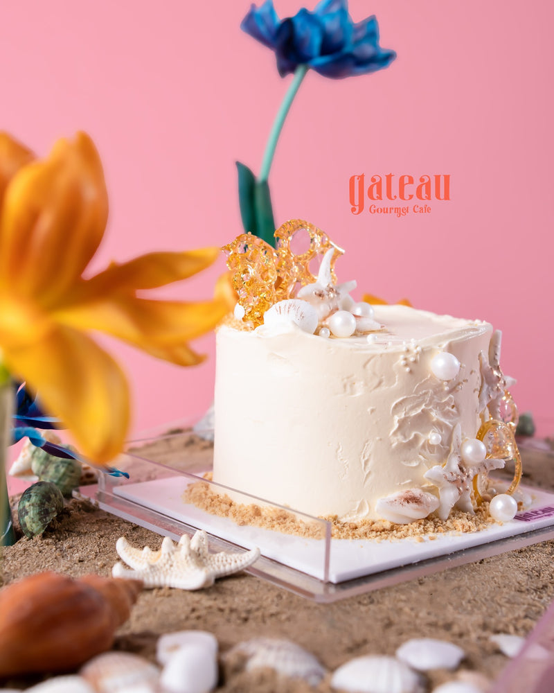 Sand and shells cake display by Gateau Gourmet Cafe featuring white frosted cake decorated with edible shells and pearls