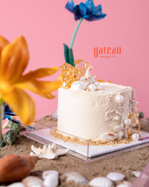 Sand and shells cake display by Gateau Gourmet Cafe featuring white frosted cake decorated with edible shells and pearls