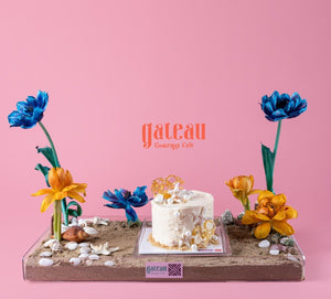 Gateau Gourmet Cafe sand and shells cake display with white cake surrounded by shells and colorful flowers on sandy base