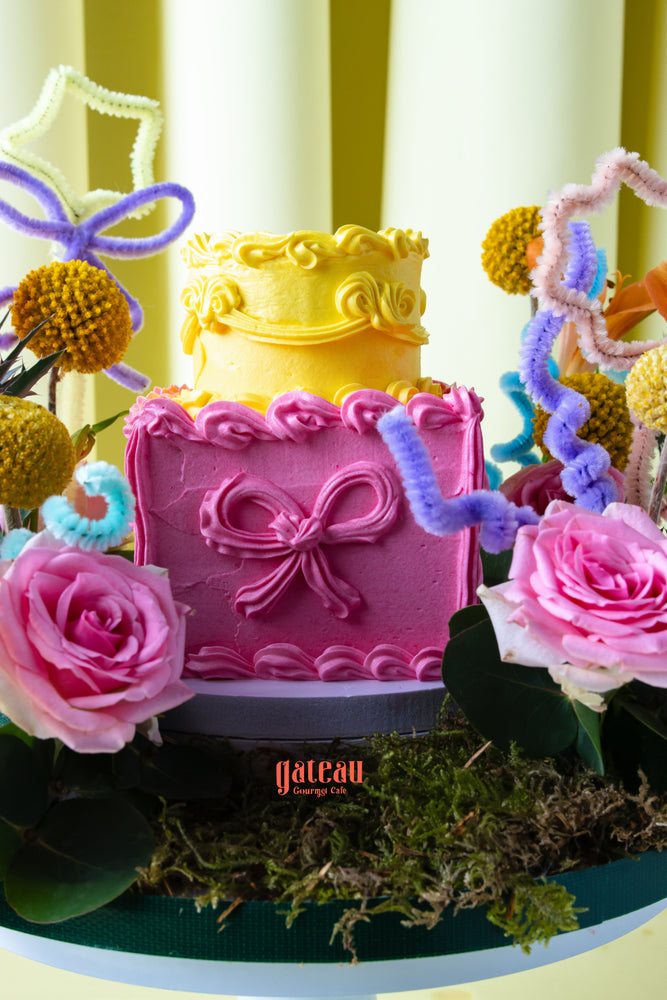 Summer buttercream flower cake by Gateau Gourmet Cafe with pink and yellow layers decorated with floral and ribbon designs