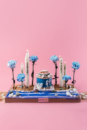 Gateau Gourmet Cafe under the sea cake arrangement with blue flowers seashells and birthday candles on pink background
