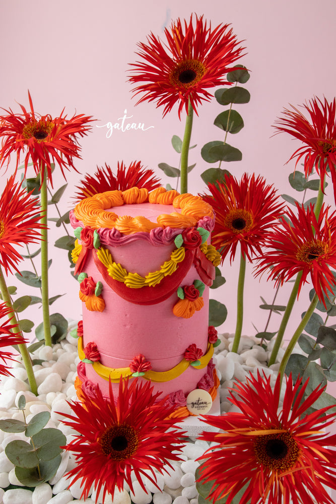 Vibrant floral cake arrangement by Gateau Gourmet Cafe featuring a pink cake decorated with colorful icing surrounded by red flowers