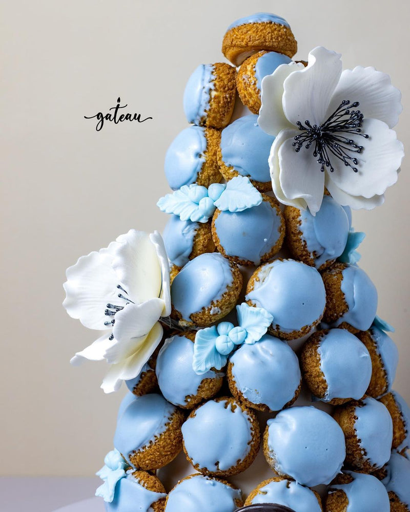 Tower of gourmet choux pastries with blue icing and white decorative flowers by Gateau Gourmet Cafe