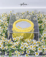 Yellow gourmet mirror cake in clear box surrounded by white daisies from Gateau Gourmet Cafe for gourmet mirror cake delivery